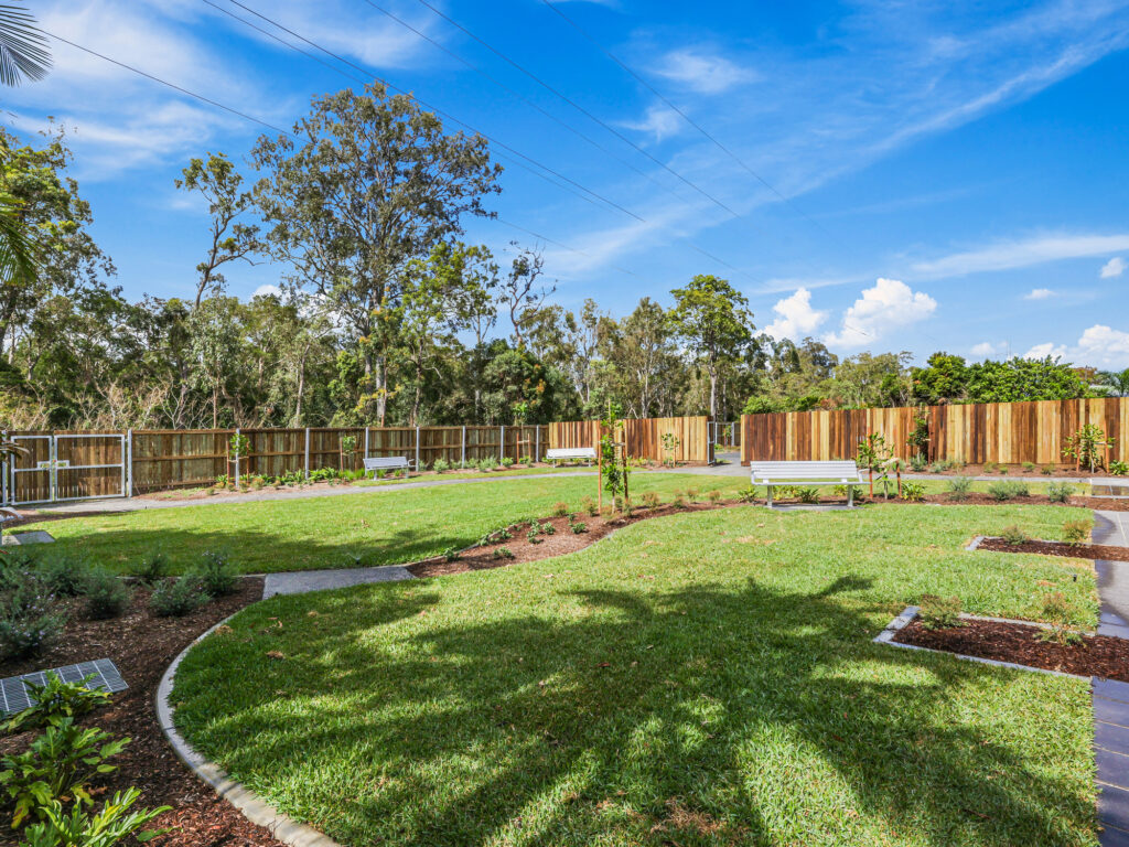 large backyard space, with multiple steel accessible permanent benches. Large backyard is framed by 6ft privacy fences, garden bed with young newly planted trees and shrubs. Backyard is flat, divided into two main sections by garden beds with plants that will grow for privacy and concrete paths across garden beds to allow for wheeled accessibility across the yard.