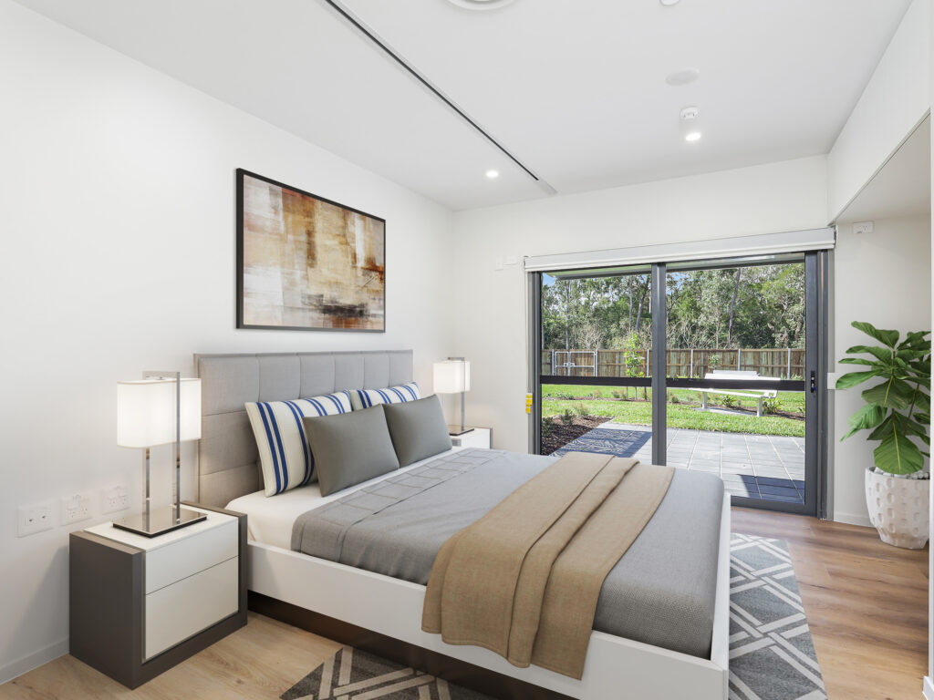 High physical support bedroom, bed in centre of photo, image taken from doorway, showing sliding glass door to large outdoor area and backyard. Tracks along ceiling for mounted ceiling hoist